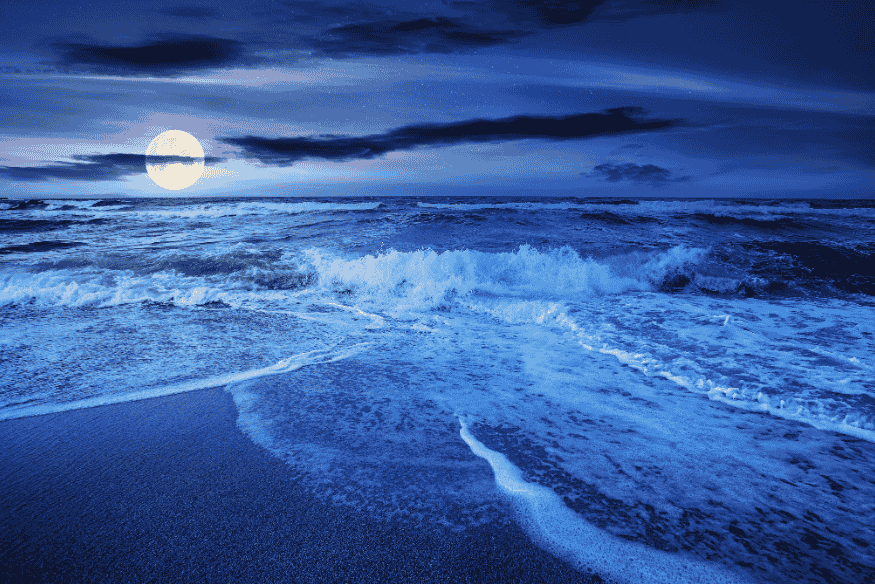 Lua cheia no horizonte sobre o oceano com ondas na praia, cenário associado às marés durante a superlua.