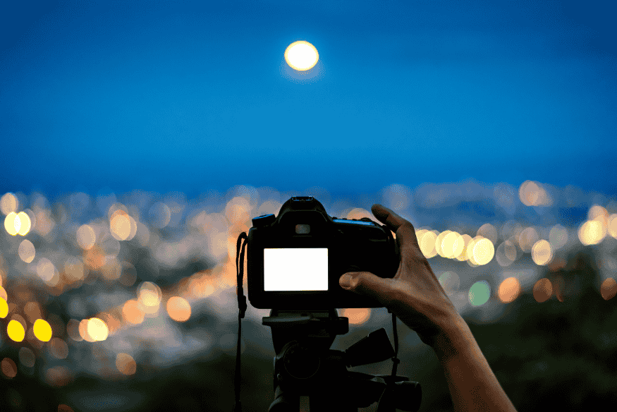 Câmera em tripé apontada para a Lua sobre cidade iluminada, exemplo de configuração para fotografar a Lua com mais estabilidade.