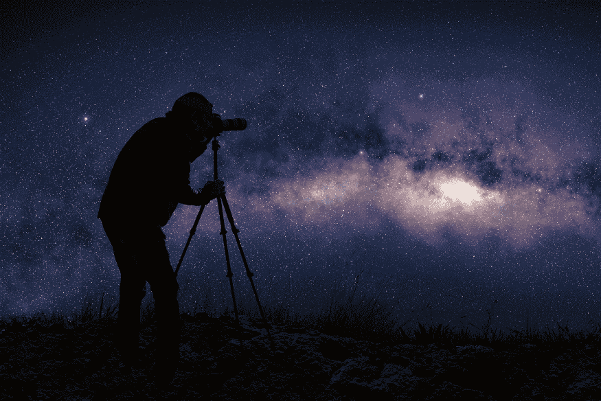 Fotógrafo com câmera em tripé registrando a Via Láctea no céu estrelado durante sessão de astrofotografia noturna.